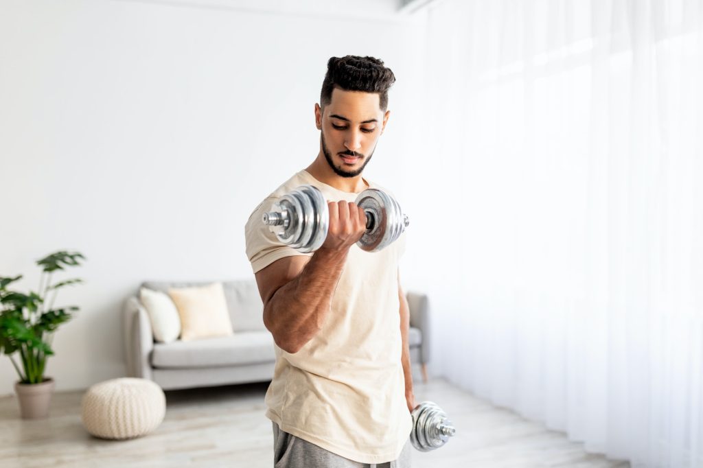 Domestic training concept. Strong young Arab man exercising with dumbbells at home