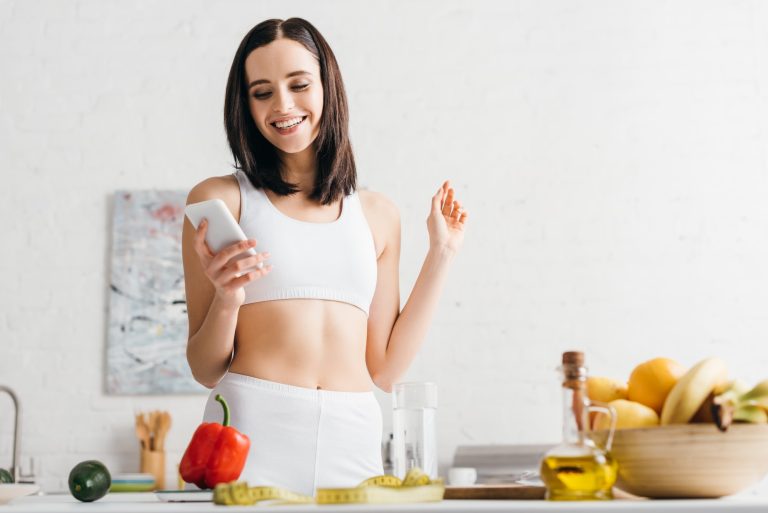 Low angle view of smiling sportswoman using smartphone near fruits, vegetables and measuring tape in