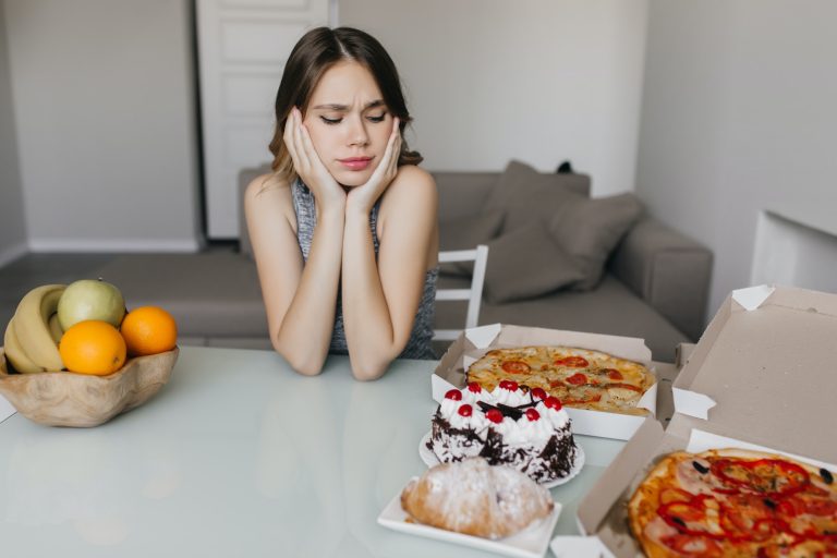 Sad curly woman looking at cake during diet. Blonde gorgeous female model posing with fruits and pi