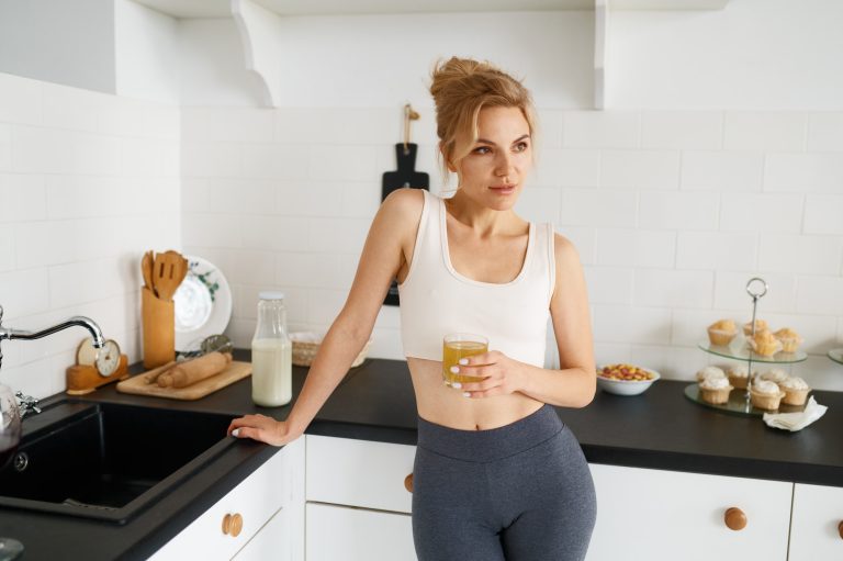 Slim woman in fitness outfit drinking fresh fruit juice on home kitchen