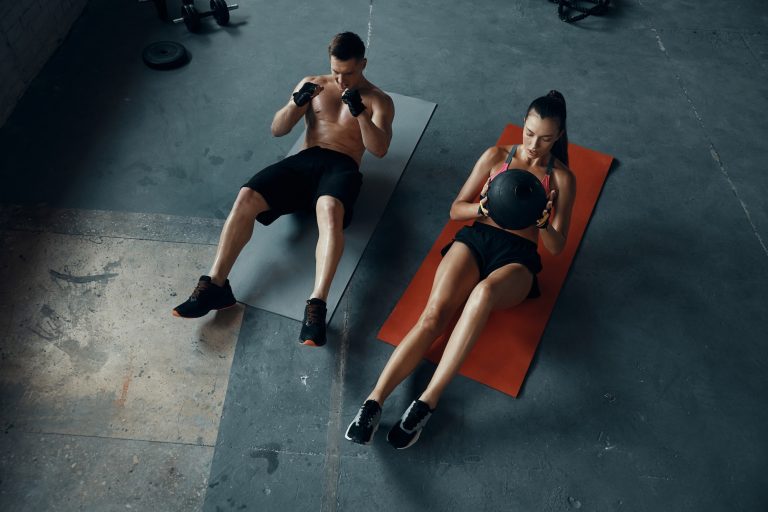 Top view of confident fit couple exercising with medicine ball while sitting on exercise mats in gym