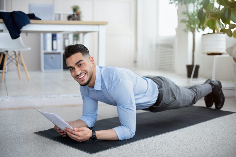 Workplace fitness. Young Arab businessman exercising on yoga mat, standing in plank and reading